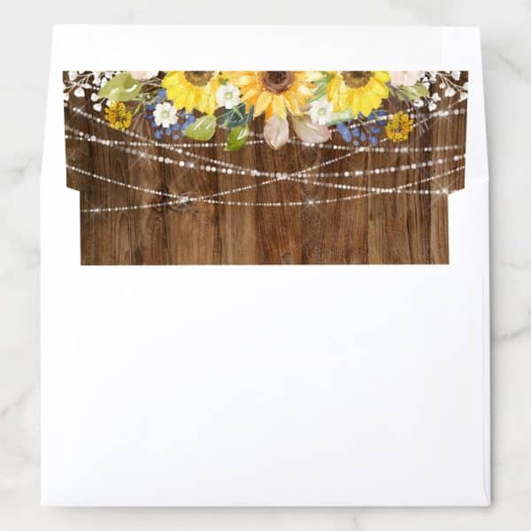 White Envelope With A Rustic Wood Background Design Featuring Sunflowers, Other Flowers, And String Lights.