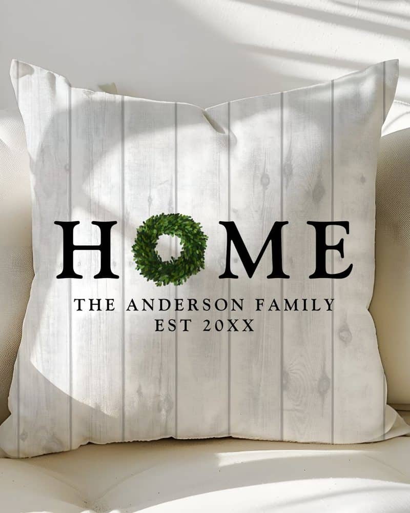 White throw pillow with the word HOME in black lettering, a green wreath replacing the letter O, and The Anderson Family Est 20XX below.