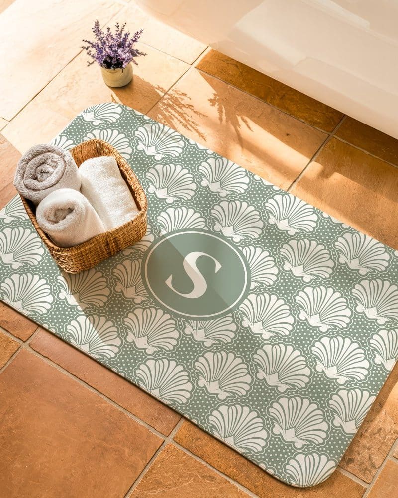 Sage green bath mat with a seashell pattern and a monogrammed S in the center.