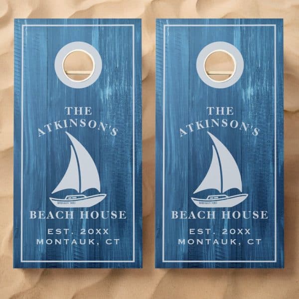 Pair Of Blue Cornhole Boards With The Atkinson'S Beach House Logo. Each Board Features A Sailboat Graphic.