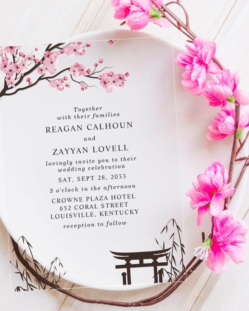 Clear wedding invitation with pink cherry blossoms and a torii gate, announcing the wedding of Reagan Calhoun and Zayyan Lovell.