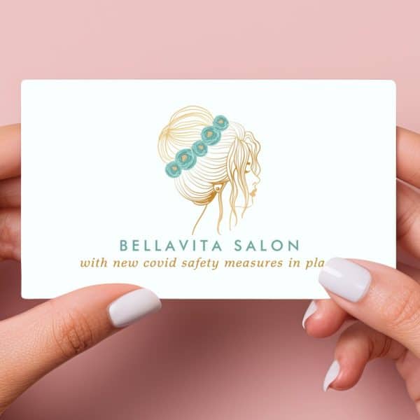 Two Hands Hold A Bellavita Salon Business Card. The Card Features A Gold Line Drawing Of A Woman'S Profile With An Updo Adorned With Teal Flowers.