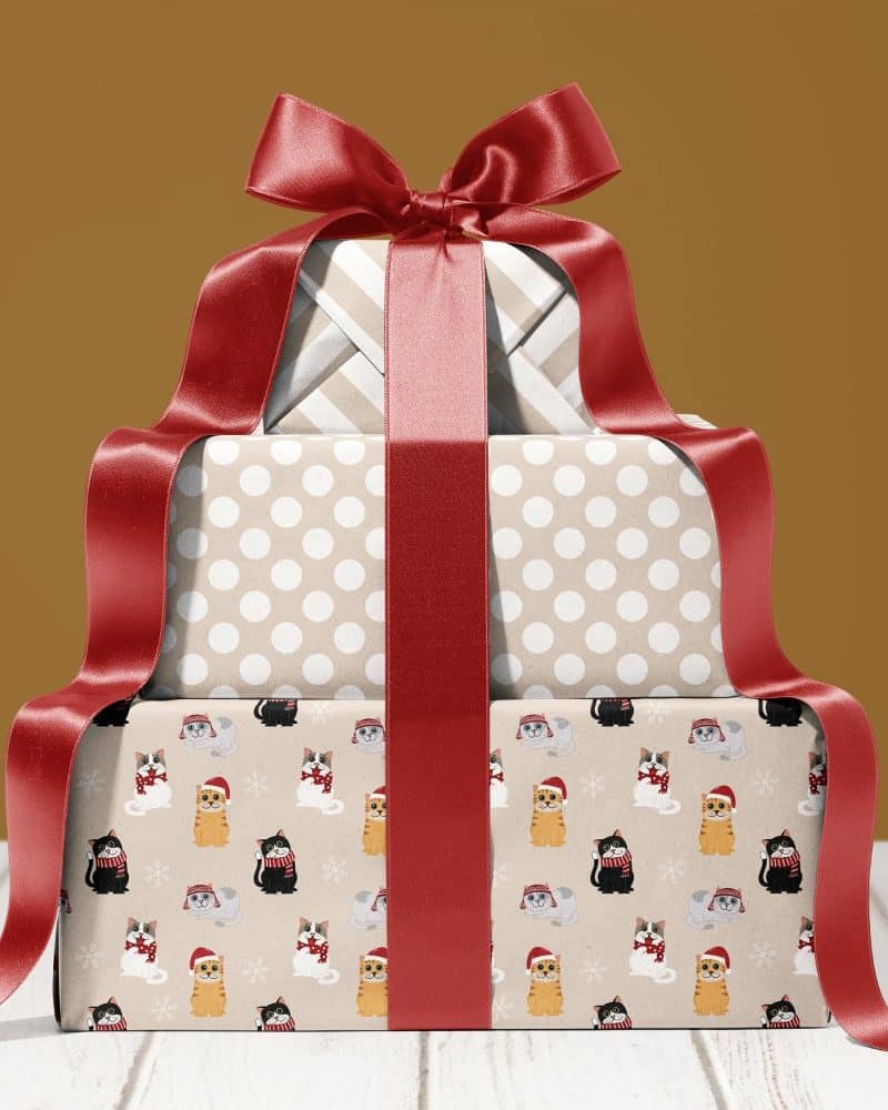 A stack of three Christmas presents tied with a large red ribbon. The bottom present features a pattern of cats wearing Santa hats and scarves.