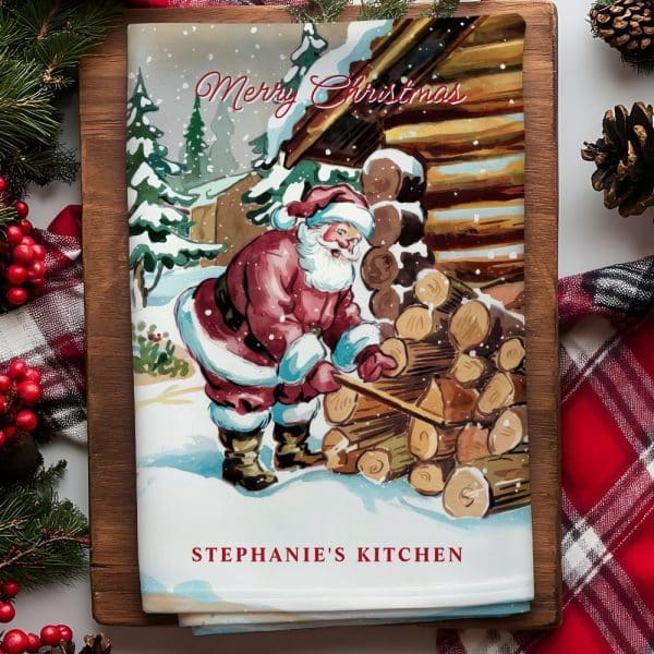 A Christmas Kitchen Towel Featuring Santa Claus Stacking Wood Next To A Snowy Cabin.