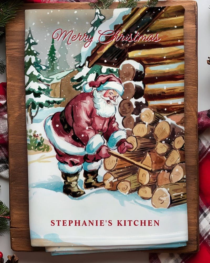A Christmas kitchen towel featuring Santa Claus stacking wood next to a snowy cabin.