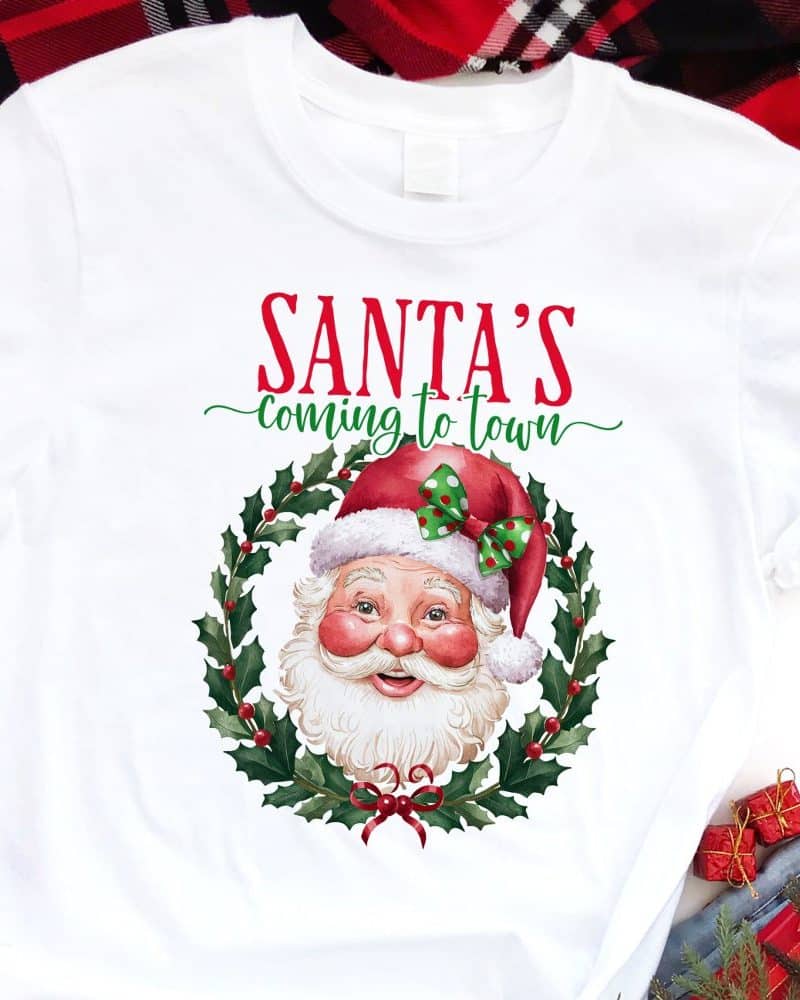 White t-shirt with a Christmas design featuring Santa's face in a holly wreath. The text Santa's Coming to Town is above the image.