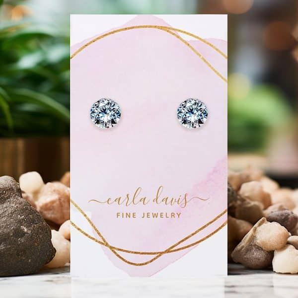 Pair Of Round, Clear Cubic Zirconia Stud Earrings Displayed On A Pink Jewelry Card.