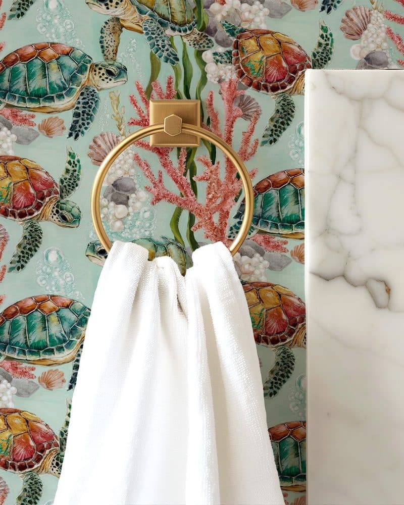 White towel hanging on a gold ring against a teal wallpaper featuring colorful sea turtles and coral.