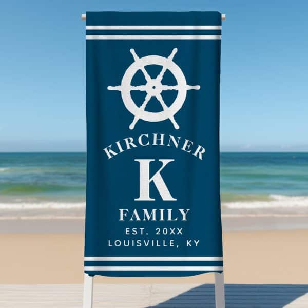 Dark Blue Beach Towel With A White Ship'S Wheel, The Letter K, And The Kirchner Family Name And Establishment Details.