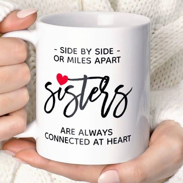 A Hand Holding A White Mug That Says Sisters Are Always Connected At Heart.