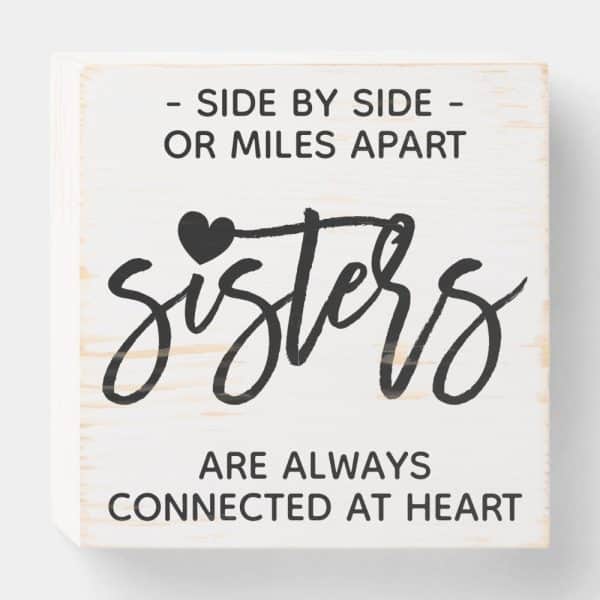 White Wooden Block Sign With The Word Sisters In Stylized Script, A Small Heart, And The Text Side By Side Or Miles Apart, Sisters Are Always Connected At Heart.