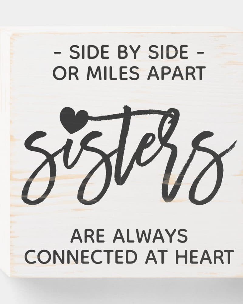 White wooden block sign with the word Sisters in stylized script, a small heart, and the text Side by side or miles apart, sisters are always connected at heart.