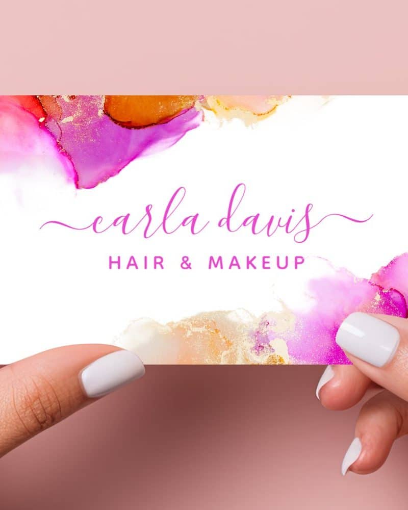Two hands hold a business card featuring an abstract alcohol ink design in pinks and golds. The name Carla Davis Hair & Makeup is displayed prominently.