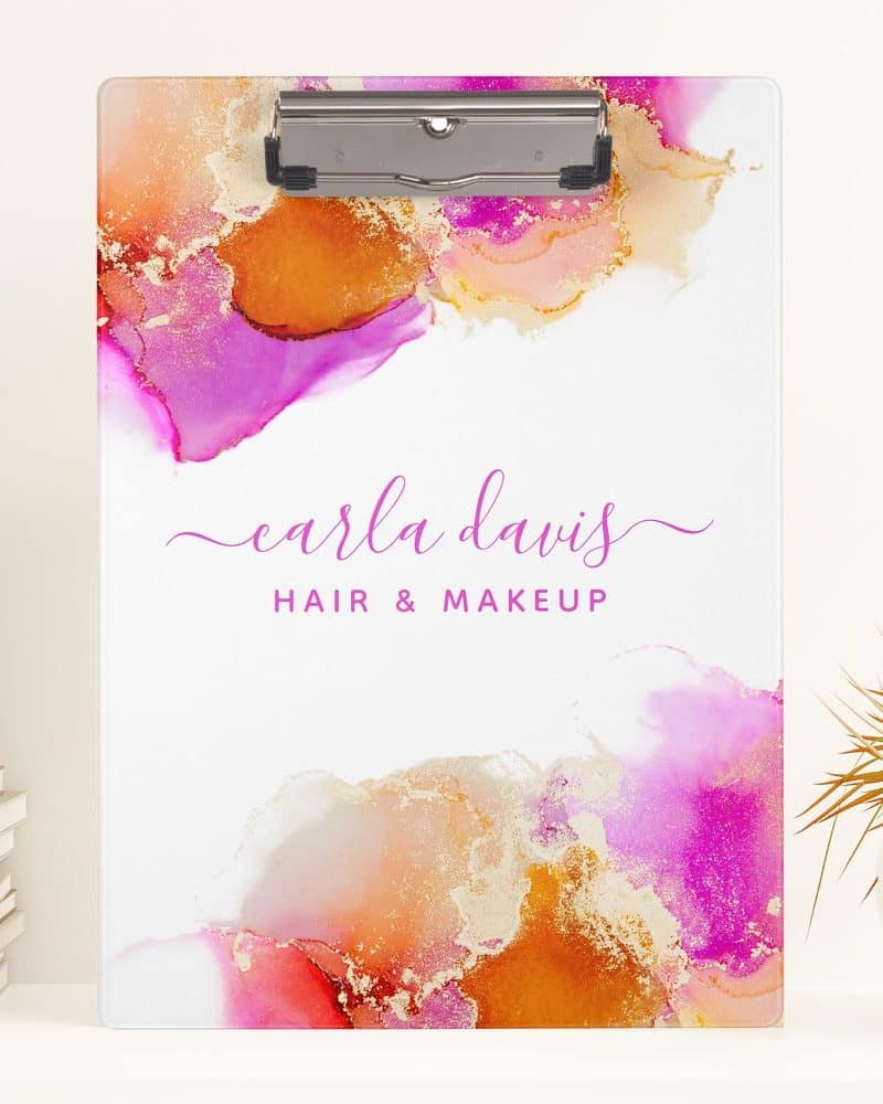 A clipboard with an abstract watercolor design in pink, orange, and gold, featuring the name Carla Davis Hair & Makeup.