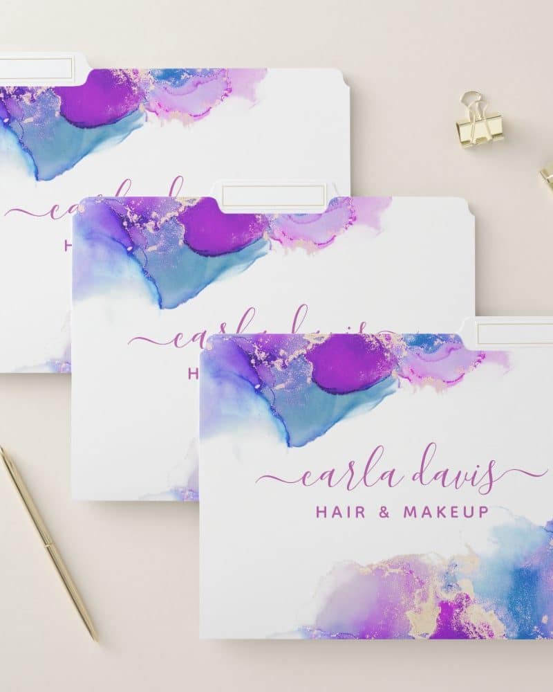 Three file folders featuring an abstract design of purple and blue watercolor paint with gold accents. The name Carla Davis Hair & Makeup is printed on the bottom folder.