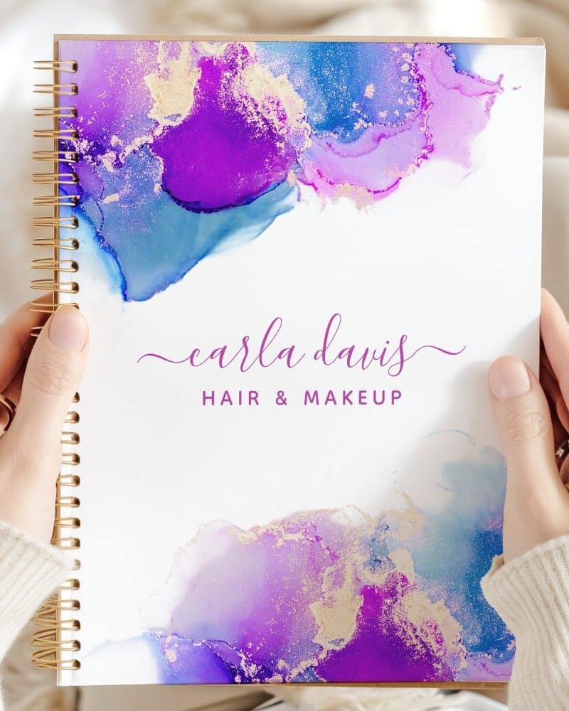Two hands hold a spiral-bound notebook featuring an abstract purple, blue, and gold design and the text Carla Davis Hair & Makeup.