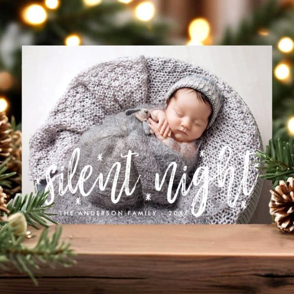Christmas Card Featuring A Sleeping Newborn Baby Nestled In A Gray Knitted Blanket. The Words Silent Night Are Overlaid On The Image.