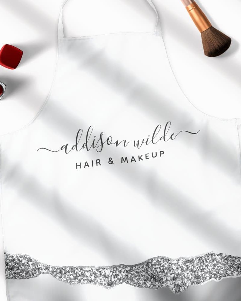 White apron with the name Addison Wilde, Hair & Makeup, and a silver glitter design at the bottom.