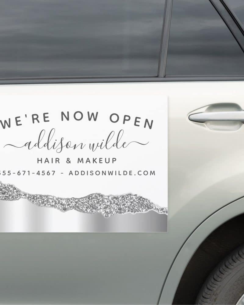 Car window sign announcing the grand opening of Addison Wilde Hair & Makeup, including phone number and website.