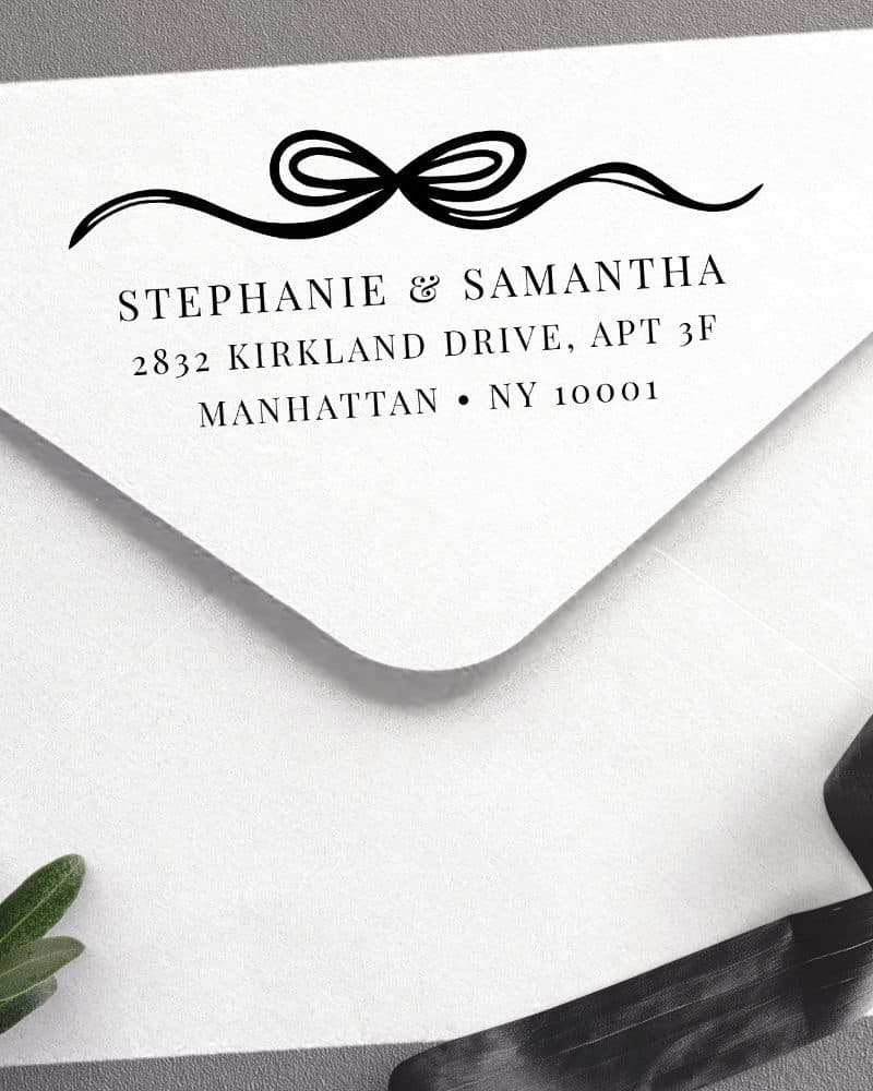 Return address stamp on a white envelope, featuring a decorative ribbon design above the names Stephanie & Samantha and their Manhattan address.