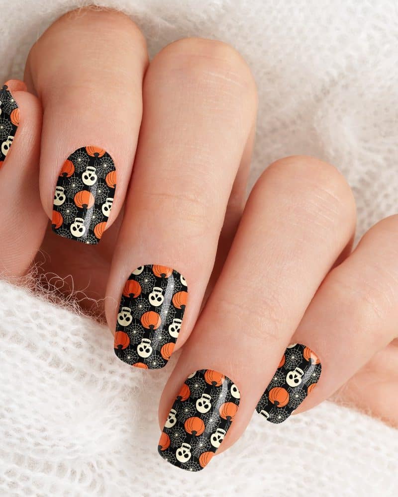 Hand with Halloween-themed nail art. The nails are decorated with a black background featuring small orange pumpkins and skulls.