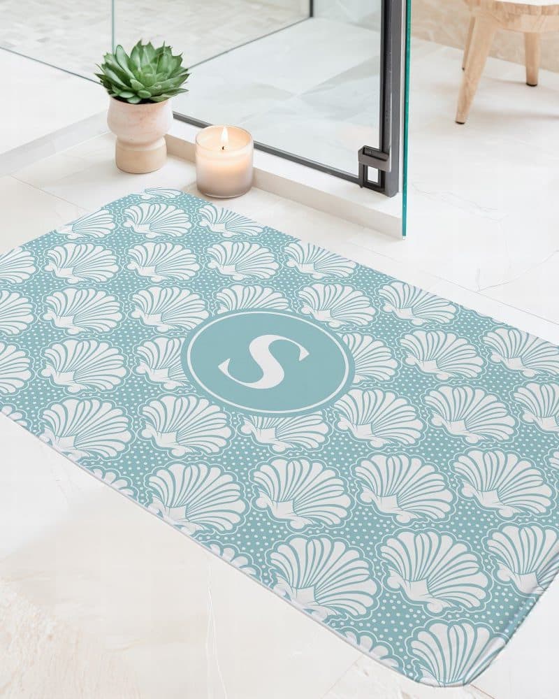 Teal bath mat with a white seashell pattern and a central teal circle containing the letter S.