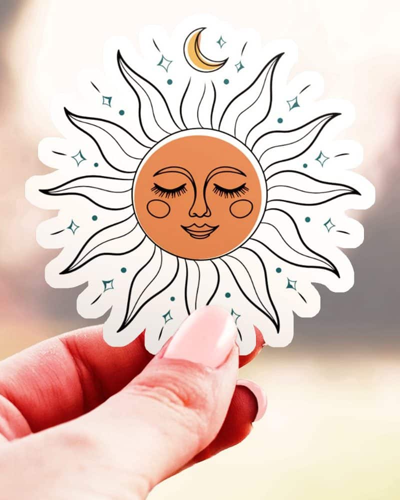 A hand holds a sticker of a stylized sun with a closed-eye face and crescent moon.