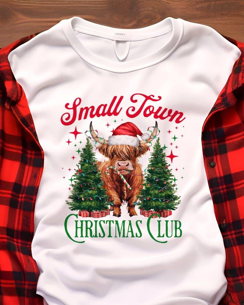 White sweatshirt with a Highland cow wearing a Santa hat, between two Christmas trees, and the text Small Town Christmas Club.