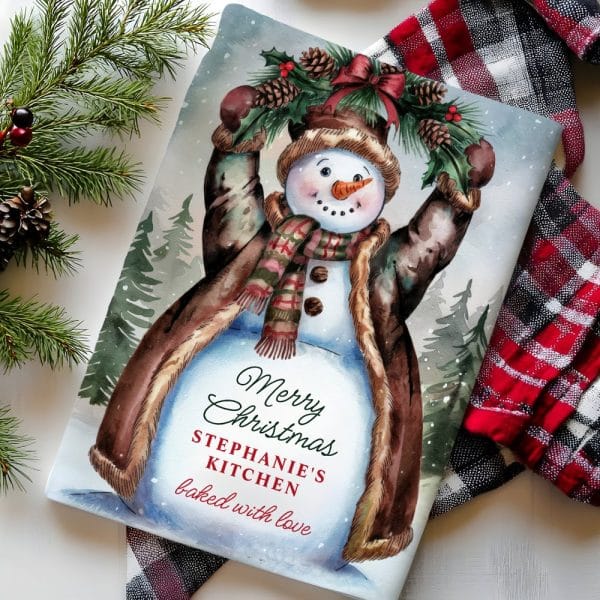 A Watercolor Painting Of A Snowman Wearing A Brown Coat, Holding A Holly And Pine Cone Wreath Above Its Head. The Text Merry Christmas, Stephanie'S Kitchen, Baked With Love Is Visible Below The Snowman.