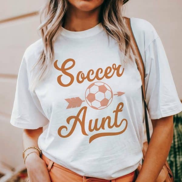 White T-Shirt With A Peach-Colored Design Reading Soccer Aunt, Featuring A Soccer Ball And Arrow.