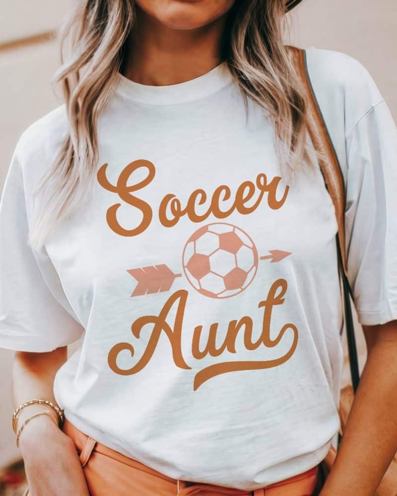 White t-shirt with a peach-colored design reading Soccer Aunt, featuring a soccer ball and arrow.