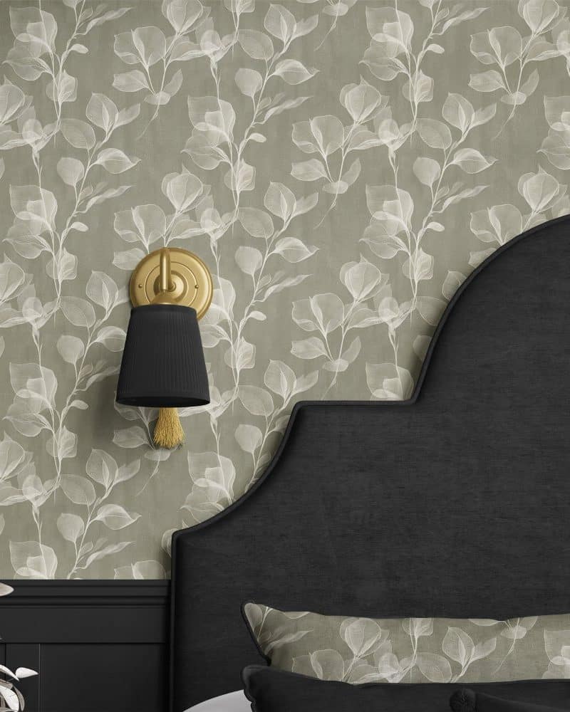A gold wall sconce with a black shade is mounted on a wall with a light green leaf patterned wallpaper. A dark headboard is partially visible.