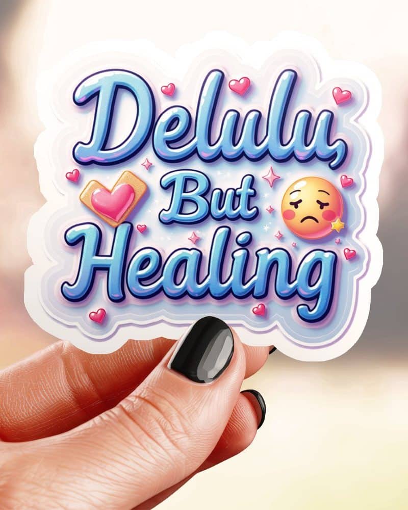 A hand holds a sticker that says Delulu But Healing in stylized blue lettering, accompanied by small hearts and an emoticon.