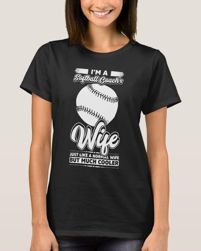 Woman wearing a black t-shirt that says Im a Softball Coach's Wife, with a softball graphic and the words Just like a normal wife but much cooler.