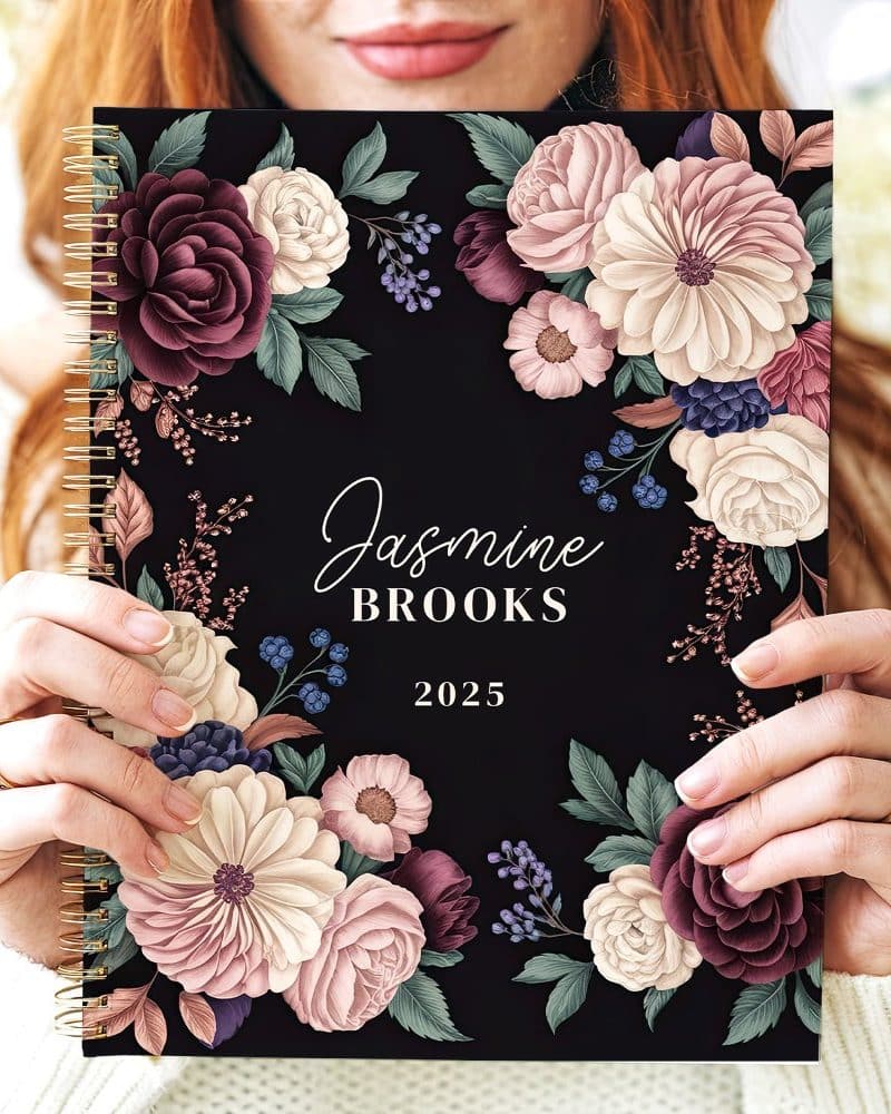 A 2025 spiral-bound planner with a dark background and a floral design featuring burgundy, blush, and cream flowers. The name Jasmine Brooks is printed on the cover.