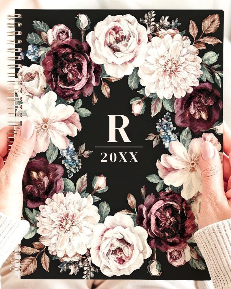 Hands hold a spiral-bound planner featuring a dark background and a floral wreath design in burgundy and cream tones. A monogrammed R and the year 20XX are centered within the wreath.