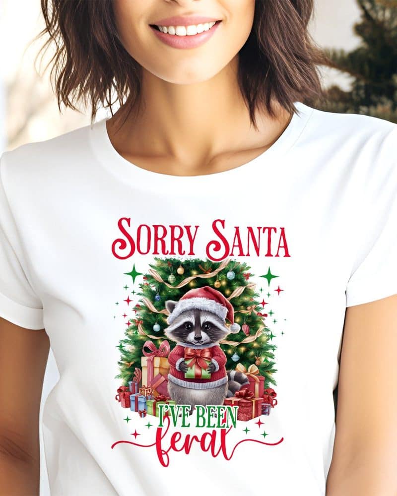 White t-shirt featuring a Christmas raccoon wearing a Santa hat and holding presents, in front of a decorated Christmas tree, with the text Sorry Santa I've Been Feral.