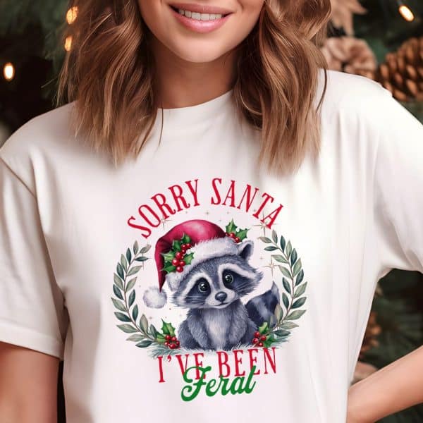 White T-Shirt Featuring A Christmas Raccoon Wearing A Santa Hat In A Wreath. The Text Reads Sorry Santa I'Ve Been Feral.