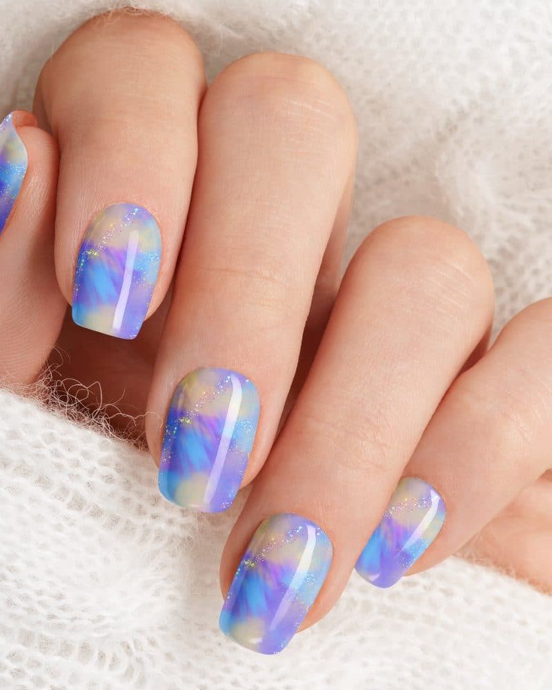 Closeup of a hand with short nails featuring a pastel purple and blue marbled nail polish design.