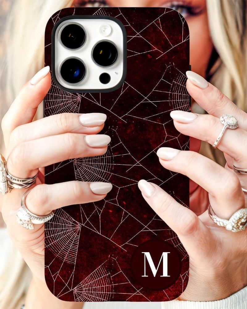 A woman's hands holding a dark red phone case with a spiderweb design and a white initial M.