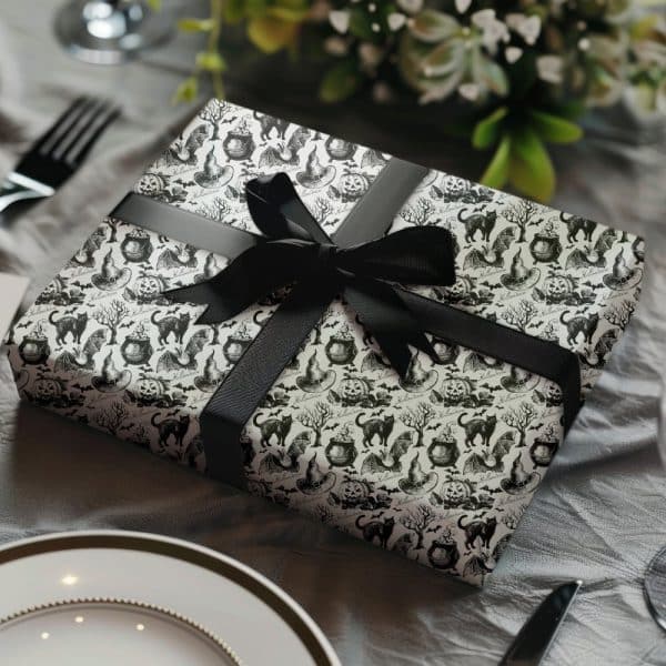 A Halloween Gift Wrapped In Black And White Patterned Paper Featuring Cats, Witches' Hats, Cauldrons, And Bats. A Black Ribbon Is Tied In A Bow On Top.