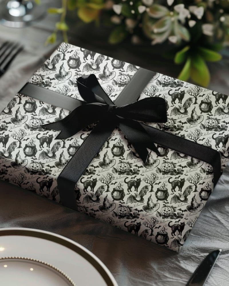 A Halloween gift wrapped in black and white patterned paper featuring cats, witches' hats, cauldrons, and bats. A black ribbon is tied in a bow on top.