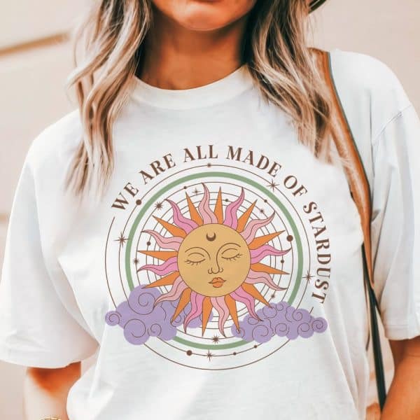 White T-Shirt Featuring A Stylized Sun With A Face, Surrounded By A Circular Design And The Text We Are All Made Of Stardust.