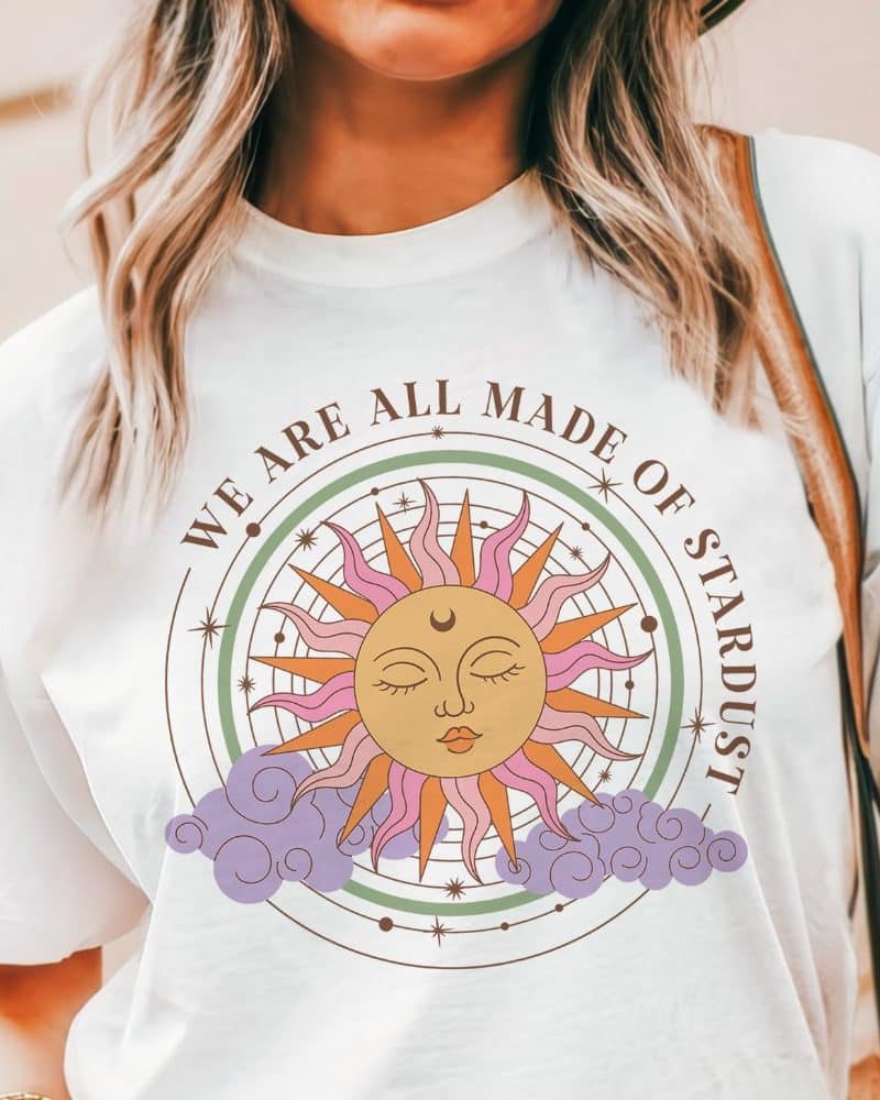 White t-shirt featuring a stylized sun with a face, surrounded by a circular design and the text We Are All Made Of Stardust.