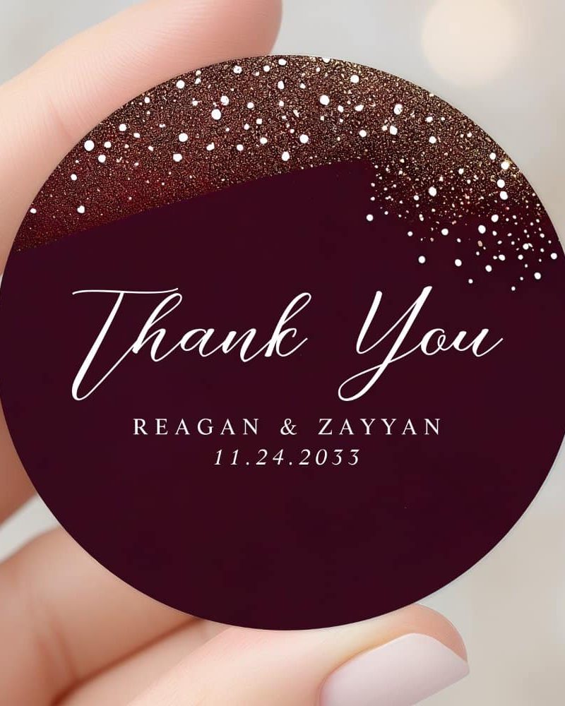 Round thank you sticker with burgundy background, gold and white glitter accents, and the names Reagan & Zayyan and date 11.24.2033.