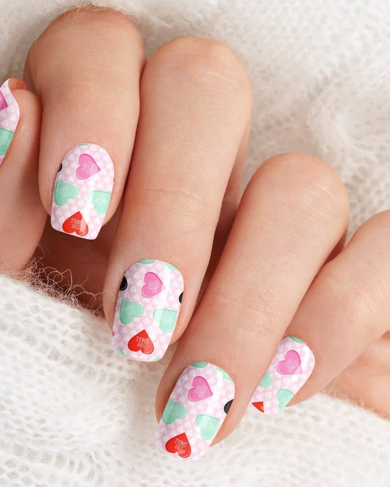 Close-up of a hand with fingernails decorated with Valentine's Day themed nail art featuring pink, red, and mint green candy hearts.