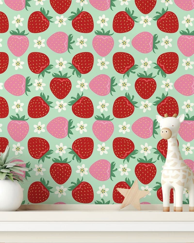 A shelf displays a wooden giraffe toy and a small plant in front of a wallpaper featuring a repeating pattern of red and pink strawberries and white flowers on a mint green background.