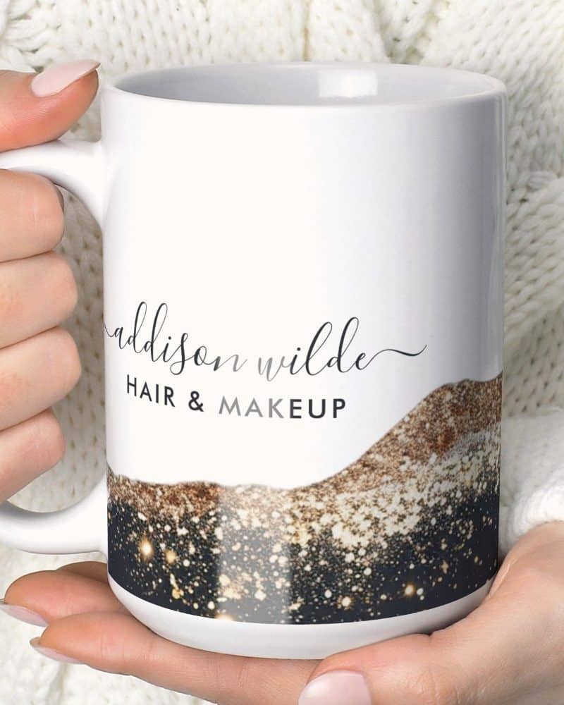 A white coffee mug with the name Addison Wilde Hair & Makeup and a sparkly gold and black design.