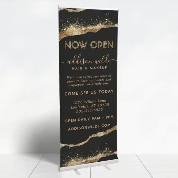 A Black And Gold &Quot;Now Open&Quot; Banner For Addison Wilde Hair &Amp; Makeup, Announcing Their Address And Hours.