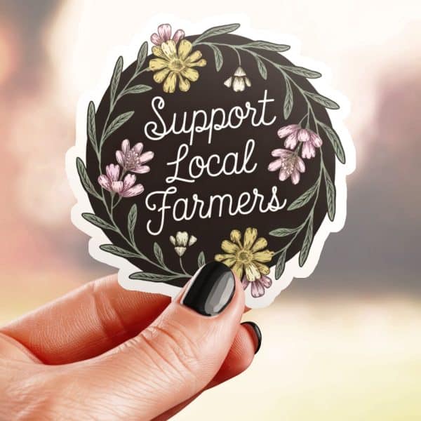A Hand Holds A Circular Sticker That Says Support Local Farmers In A Floral Wreath.